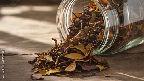 Dried herbal tea leaves spilling from glass jar onto rustic wooden surface with wooden table still life close up ingredients background collection