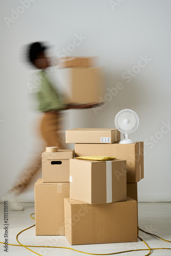 Vertical shot of young adult woman carrying cardboard boxes past stacked parcels, suggesting packing, moving, shipping workflow. Ideal for e commerce, delivery, relocation marketing