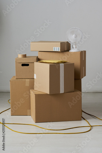 Vertical shot of stacked cardboard boxes, parcel mailer, packing tape, fan, extension cord suggesting home relocation, storage, shipping logistics. For moving service, delivery, decluttering ads
