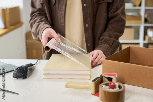 Young adult worker wrapping books with plastic film at packing station, preparing inventory for shipment. Image supporting e commerce fulfillment, order processing, retail logistics