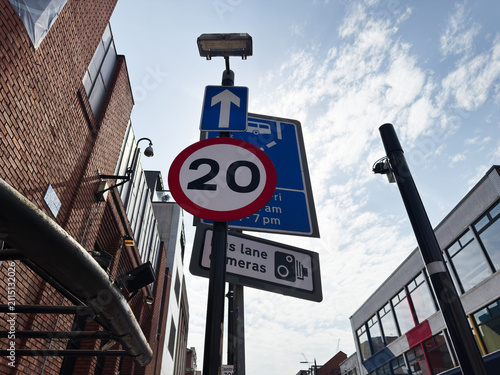 Traffic sign with 20 speed limit and one way arrow on a city street in Manchester, UK showing bus lane camera and urban buildings