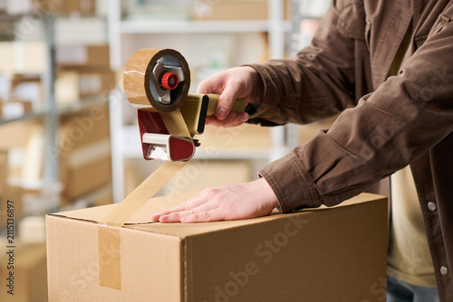 Close up of young adult worker sealing cardboard box with packing tape in warehouse, preparing shipment for order fulfillment. Ideal for e commerce, logistics, delivery marketing