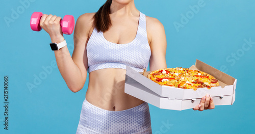 Anonymous athlete holding pizza box while pumping muscles with dumbbell to reject junk food isolated over blue background