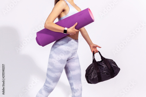 Anonymous person in white sportswear holding bag and fitness mat to hurry for gym workout isolated over gray background with copy space