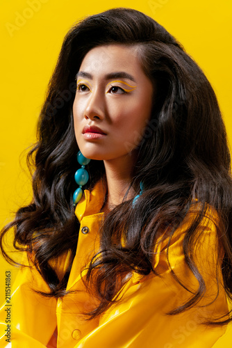 Asian woman in yellow raincoat wearing blue stone earrings and creative eyeliner looking away against bright background