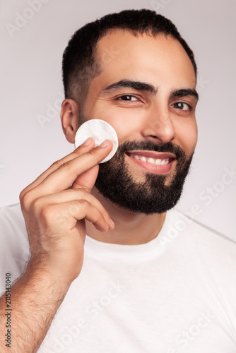 Smiling man applying skincare lotion with cotton pad wiping bearded face for hygiene wearing casual white t-shirt posing isolated over gray background