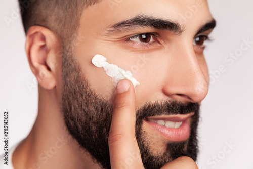 Close up of man spreading white facial cream with finger on cheek area for intensive skin hydration during grooming session isolated over gray background