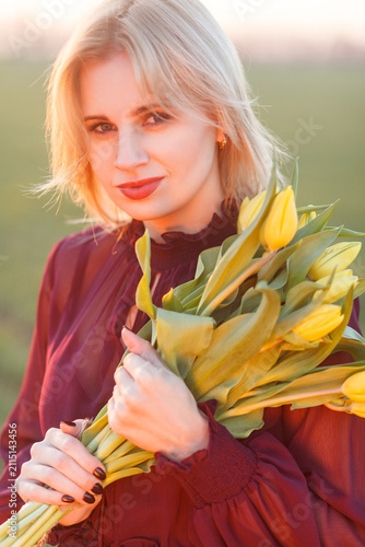 Blonde woman holding yellow tulips near her face while looking at camera in field during sunset with soft lens flare