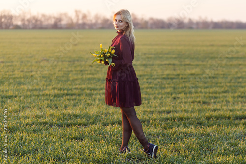 Woman with short blonde hair in tiered burgundy ruffled dress posing in green field during sunset while holding a bouquet of yellow tulips in hands and looking at camera