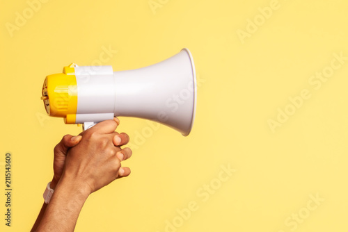Profile portrait of astonished worker wearing blue overalls and red hat holding megaphone in hands and looking at camera with widely open mouth. Indoor studio shot isolated on yellow background.