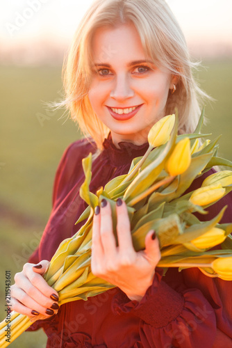 Happy woman holding yellow tulips looking at camera with radiant smile standing in green spring field at sunset