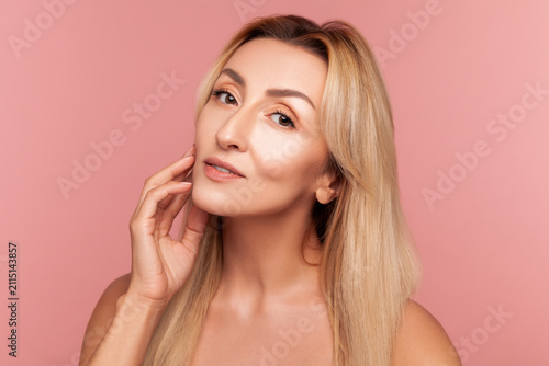 Beautiful adult woman touching her smooth face skin gently for aesthetic dermatology and rejuvenation campaign isolated on pink background