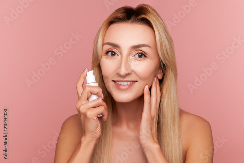 Smiling woman holding white serum bottle for face hydration and anti-aging treatment during daily cosmetic beauty routine isolated on pink background