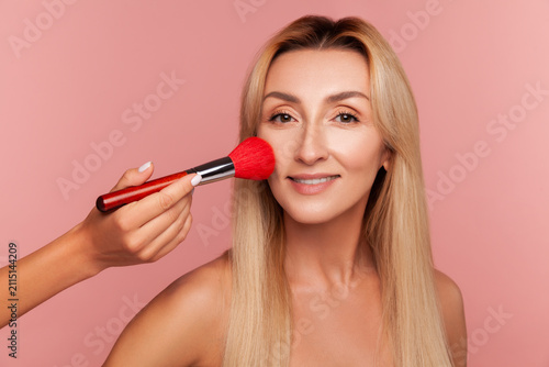 Blonde female model getting makeup with large red brush professional artist enhancing skin tone and beauty isolated on pink background