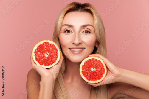 Happy woman holding two grapefruit halves for skin health and vitamin C infusion beauty procedure isolated on pink background