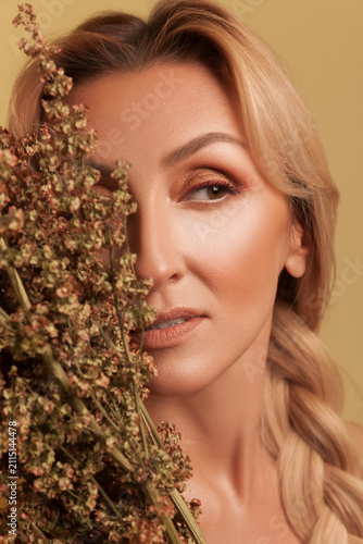 Beautiful female face partially hidden by dried plants while looking away showing bronze eye makeup isolated over yellow background