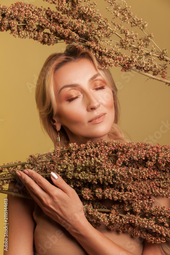Serene blonde lady holding dried sorghum plants with eyes closed enjoying autumn atmosphere isolated over yellow background