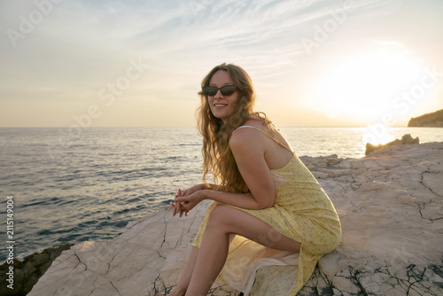 Принти на полотні Young woman in yellow dress sitting on rocky sea coast enjoying beautiful sunset