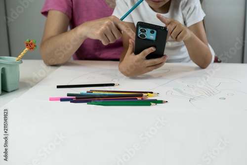 фотографія A young left-handed student uses a mobile phone at a table with drawing paper and pencils while studying with a parent
