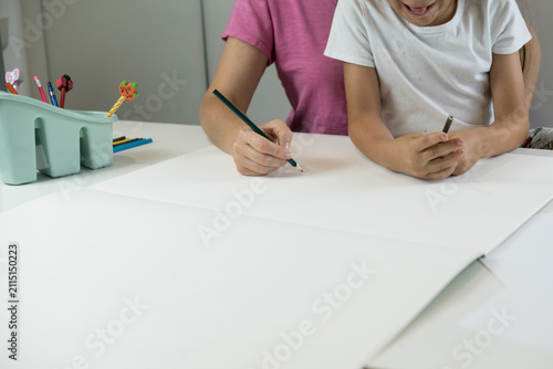 Mother holding pencil on paper guiding left handed child sitting together at white desk during home study.