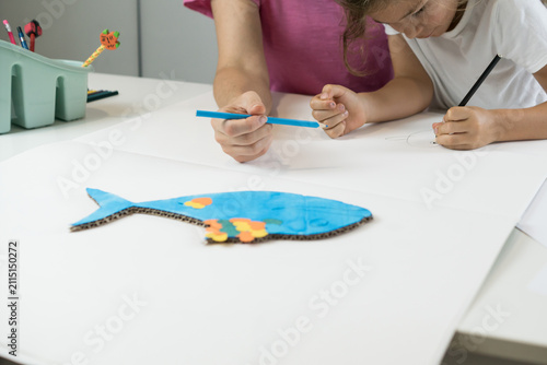 Fotografie Close-up of a left-handed child holding a blue pencil and creating a colorful fish craft on a white paper surface during art class