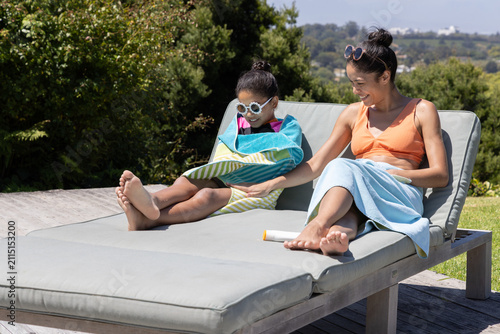 Woman and child sitting on cushioned lounge on wood deck in towels applying sunscreen
