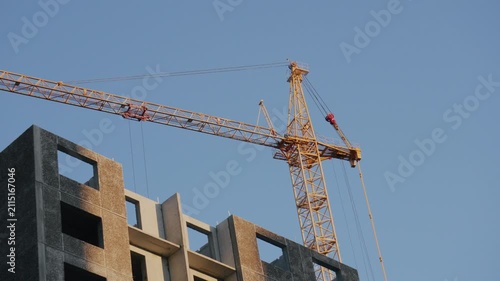 Two tall cranes stand in an urban area during the day. The cranes are positioned against a clear blue sky
