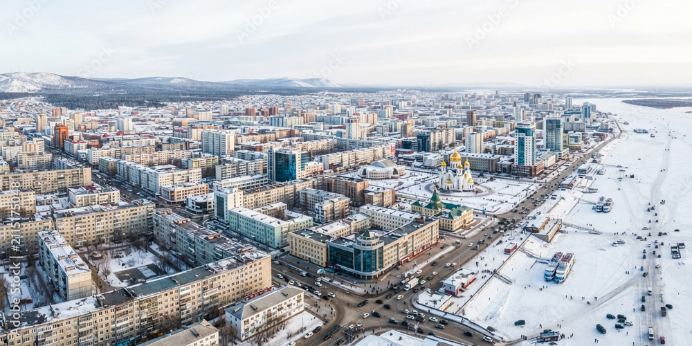 Fototapeta premium Aerial view of a snowy winter city adjacent to a frozen river and distant hills under a soft winter sky