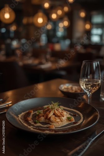 Seared scallops in a rich sauce, garnished with fresh microgreens and served on a dark plate alongside a glass of white wine.
