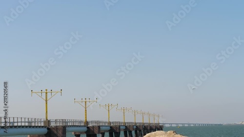 Runway approach lighting system pier extending over the calm green sea under a clear blue sky, featuring a distant commercial airplane descending, wide static shot.