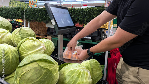 Customer Sticking Price Barcode Sticker On Bag Of Carrots On Supermarket Scale Near Cabbages