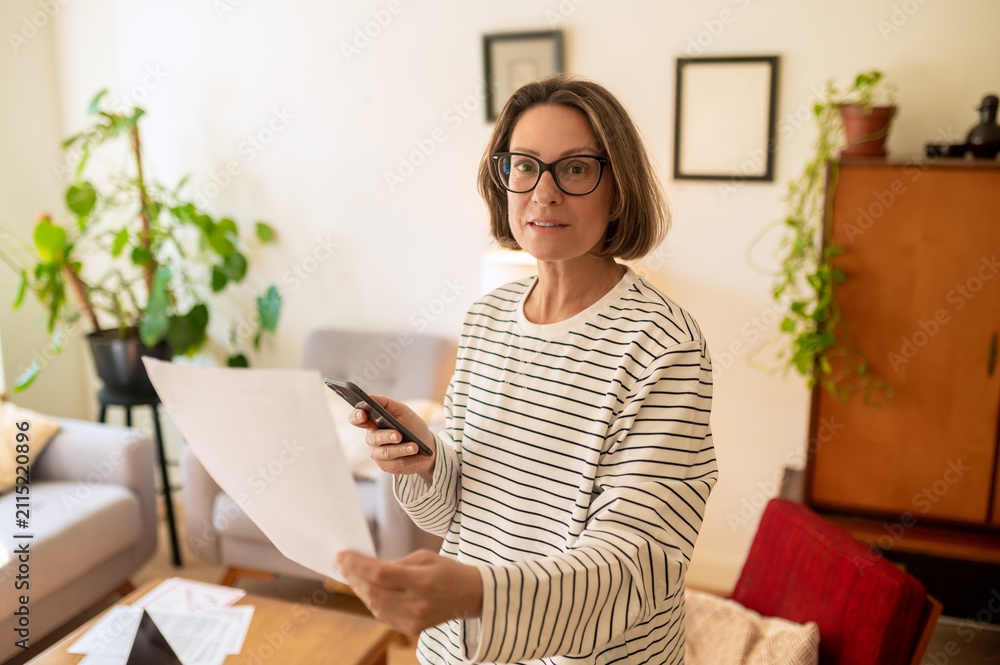 Obraz premium Middle aged woman standing with paper and smartphone