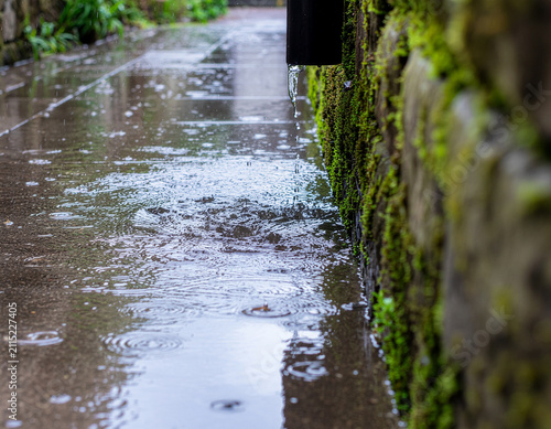 low angle shot of rainwater pouring out of a drain pipe onto a wet stone walkway lined with a moss covered wall