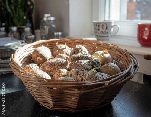 ​a woven wicker basket filled with sprouting seed potatoes placed on a dark kitchen counter near a sunny windowsill
