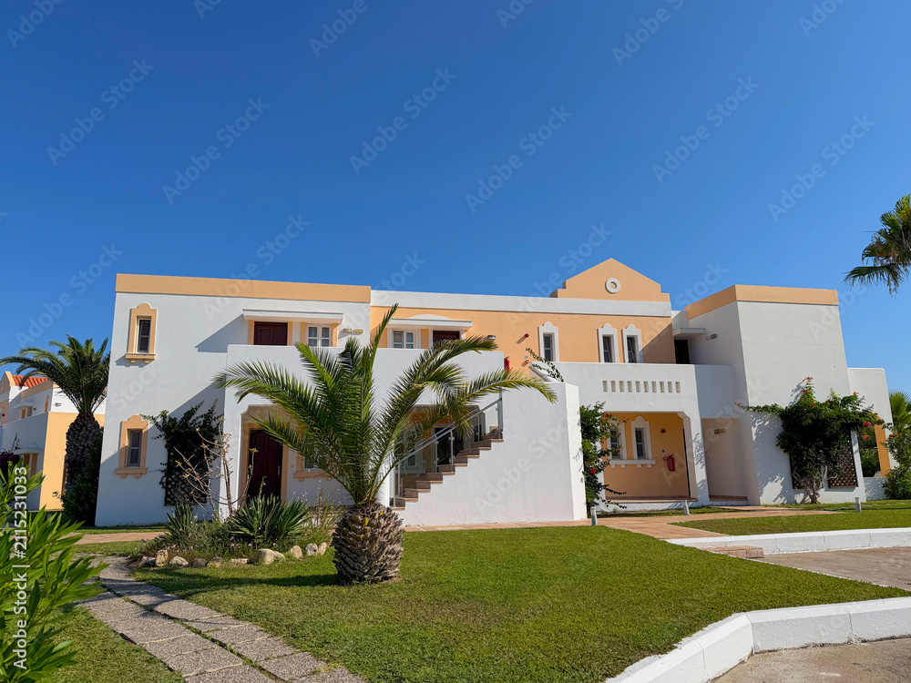 Fototapeta premium Mediterranean white and peach stucco resort villa with palm trees and lush green lawn under a bright clear blue sky on a sunny day