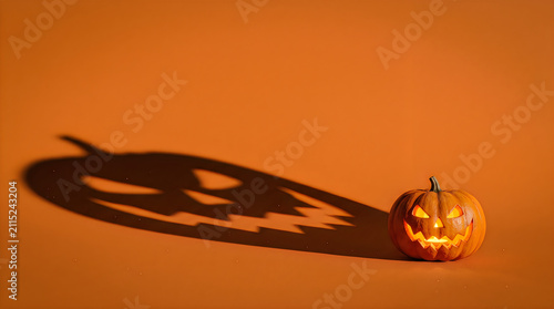 Small carved pumpkin with illuminated features rests on a dark surface, casting a long, eerie shadow across a bright orange background.