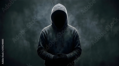Hooded figure stands in darkness, with hands clasped and face obscured, creating a mysterious and anonymous silhouette.