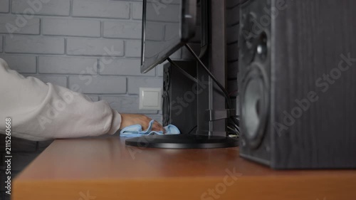 Person cleaning a dusty wooden computer desk with a microfiber cloth. Person's hand cleaning a brown wooden office desk with a blue microfiber cloth, wiping dust around a computer monitor and speakers