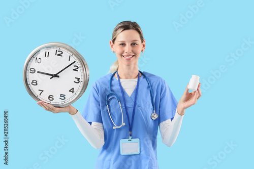 Female nurse in blue scrubs holding a large clock and a medicine bottle, Heal...