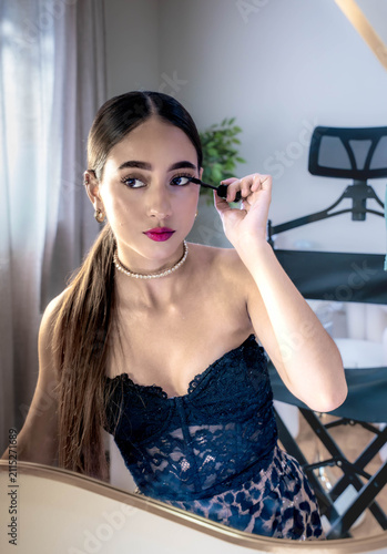 Young woman carefully applies mascara beside mirror in bright bedroom light, wearing blue lace dress, pearl choker, and gold earrings, creating elegant beauty routine scene with soft window glow and