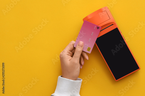 Female hand with credit card and payment terminal on yellow background
