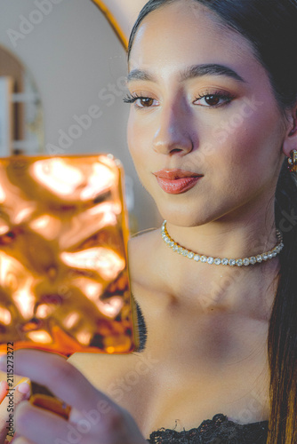 Close up portrait of young woman checking makeup in gold hand mirror indoors, with soft warm lighting, dark hair, glossy lips, sparkling choker, lace top, and calm focused expression