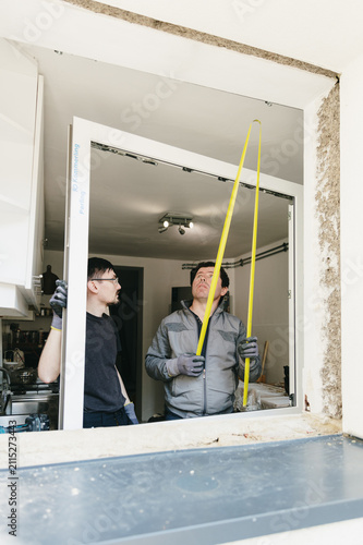 Vertical portrait of two workers measuring a window frame with a yellow tape measure.