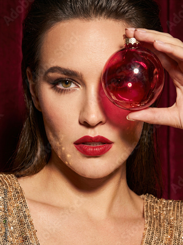 Beautiful elegant woman with red lipstick and gold sequin dress holding a red Christmas ornament over her eye in a festive studio setting