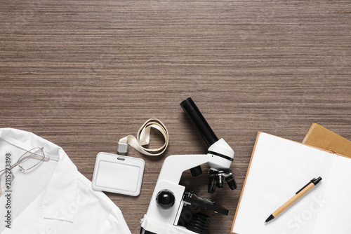 Modern microscope with lab coat, notebook and name tag on wooden background