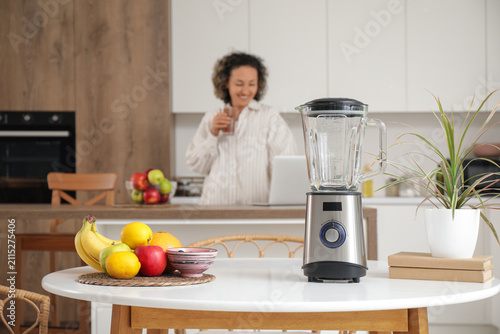 Blender with fruits on table of mature woman using laptop in kitchen, closeup