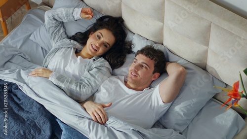 Konstfotografi Happy Caucasian couple man woman lying on bed at home bedroom talking together c