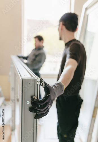 A vertical portrait of a worker helping to hold a window frame while a craftsman takes measurements with a tape measure in a bedroom.