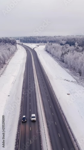 Aerial shot of a four lane highway during wintertime. Cloudy sky. Snowy forest and ground. Cold nordic weather. Vehicles driving. Drone slowly zooming out. Vertical video