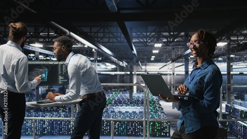 Canvastavla A happy woman in data center performs maintenance tasks on laptop, ensuring optimal performance; an engineering team reviews diagnostics and applying patches to server room, improving stability